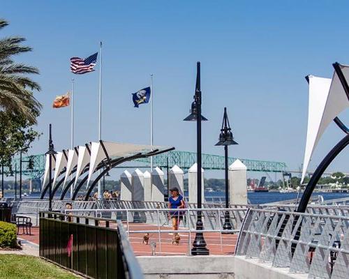 Photo of Jacksonville's 南岸河边漫步 during a sunny day. Brick sidewalk alongside the river with 钢护栏. Canopies overhang the sidewalk providing shade.